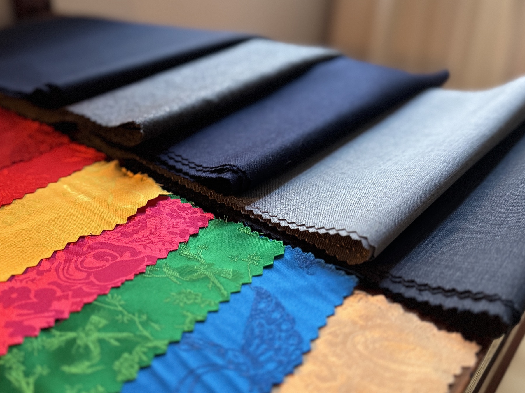 Folded suiting cloths beside jacquard linings in red, gold, magenta, green, blue and bronze, each with a pinked edge.